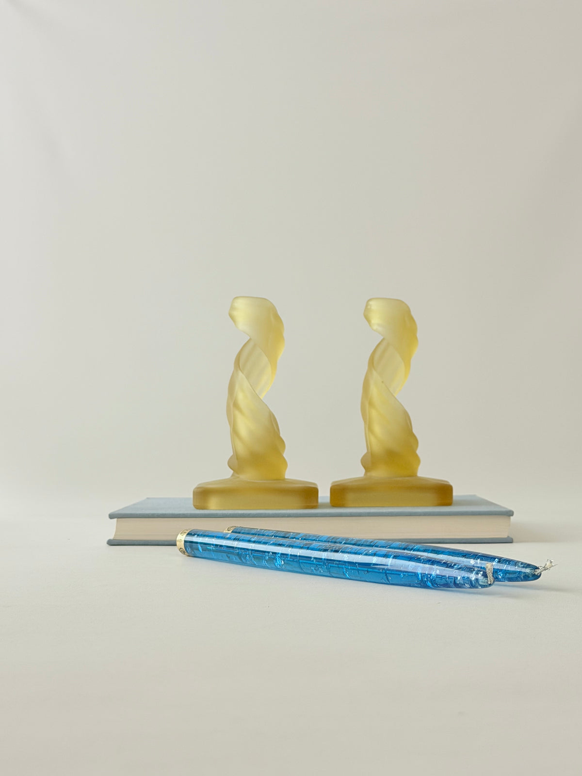 A pair of  yellow frosted glass twisted glass candleholders, displayed on a book with tall blue lucite candles.